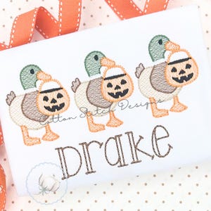 Può includere: Una superficie in tessuto bianco con un ricamo raffigurante tre germani reali, ognuno con una testa di zucca intagliata. Il testo "Cotton Stitch Designs" è visibile e il nome "Drake" è ricamato in marrone sotto le anatre.