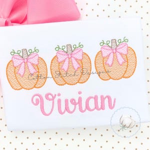 May include: A white shirt featuring an embroidered design of three orange pumpkins, each adorned with a light pink bow and green stems. Below the pumpkins, the name Vivian is embroidered in a cursive pink font. A pink bow sits at the top.