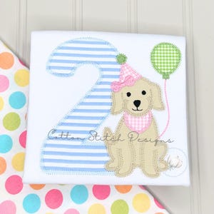 May include: White t-shirt with a light blue striped number 2 applique. A tan puppy wearing a pink party hat and bib is next to the number. A green gingham balloon is held by the puppy. The text "Cotton Stitch Designs" is on the shirt.