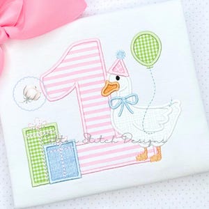 Peut inclure: T-shirt blanc avec un motif brodé représentant le chiffre 1 à rayures roses, une oie portant un chapeau de fête et un nœud papillon, un ballon et des cadeaux emballés. Le texte "Cotton Stitch Designs" est également présent.
