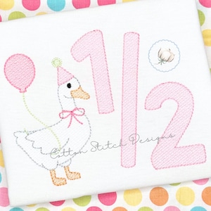 May include: White fabric with a pink embroidered design featuring a goose wearing a party hat and holding a balloon. The numbers "1/2" are also embroidered in pink, along with a small cotton illustration. The text "Cotton Stitch Designs" is also embroidered.