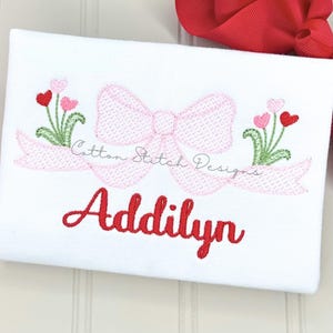 May include: White fabric with embroidered design. A pink bow and banner with the text "Cotton Stitch Designs" and "Addilyn" in red script. Features embroidered flowers with heart-shaped petals.
