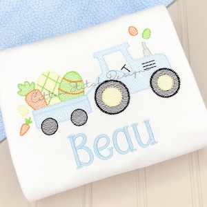 May include: White fabric with a light blue tractor design carrying Easter eggs and carrots. The tractor has black wheels and the name "Beau" embroidered in light blue. The design includes orange, green, and yellow accents.