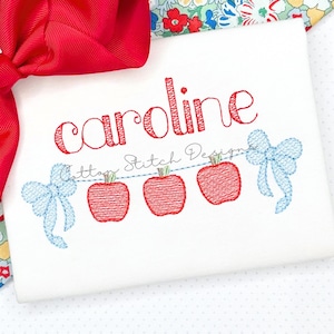 May include: White t-shirt with the name "Caroline" embroidered in red script. Below the name are three red apple designs strung on a blue dotted line, with blue bow accents on each end. The words "Cotton Stitch Design" are below the name.