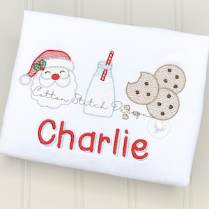 May include: White t-shirt with an embroidered design featuring Santa Claus, a milk bottle with a red straw, and three cookies. The name "Charlie" is embroidered in red with a blue outline. The text "Cotton Stitch Designs" is also embroidered.