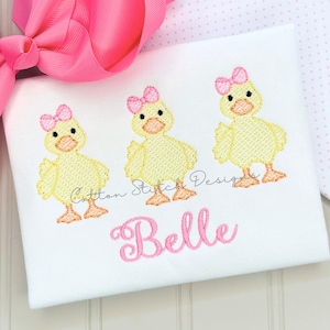May include: White baby shirt embroidered with three yellow ducklings wearing pink bows. The name "Belle" is embroidered in pink script below the ducks. A large pink bow is in the upper left corner.