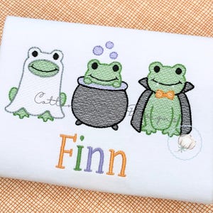 May include: A white fabric item featuring an embroidered design of three green frogs. One frog wears a white ghost costume, one sits in a black cauldron with purple bubbles, and one wears a black cape with an orange bow tie. The name Finn is embroidered below in orange, green, and purple letters.