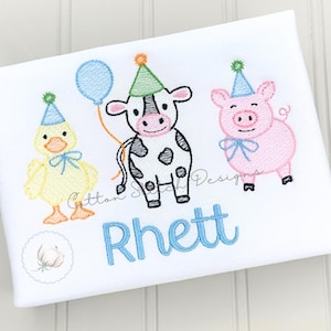Puede incluir: Camiseta blanca con animales de dibujos animados bordados: un pato amarillo, una vaca blanca y negra y un cerdo rosa, todos con gorros de fiesta. Un globo azul y el nombre "Rhett" también están bordados en la camiseta.