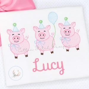 May include: White embroidered fabric featuring three pink pigs wearing party hats and holding a blue balloon. The name "Lucy" is embroidered in pink script below the pigs. A pink bow is visible in the upper left corner.