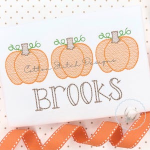 May include: A white fabric surface featuring an embroidered design of three orange pumpkins with green vines and grey stems. Below the pumpkins, the name BROOKS is embroidered in grey thread. An orange ribbon with white stitching is at the bottom.
