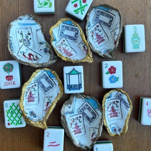 May include: A selection of white Mahjong tiles and oyster shells. The shells are painted with Asian-inspired designs in red, blue, and gold. The tiles feature various symbols and numbers, including a "JOKER" tile.