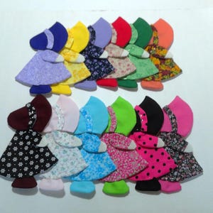May include: A collection of fourteen fabric doll ornaments arranged in two rows. Each doll features a bonnet and a dress in various colors and patterns, including solid, floral, and polka dot prints, with white fabric hands and feet.