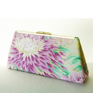 Pu&ograve; includere: Una pochette floreale con chiusura dorata. Il tessuto presenta un grande fiore viola e bianco con foglie verdi su uno sfondo crema.