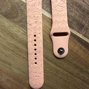 Puede incluir: Dos correas de reloj de color rosa claro. Una correa presenta una serie de caras de animales de dibujos animados y la palabra "Stay" repetida. La otra correa tiene la palabra "Stay" repetida y un diseño circular. Ambas correas están hechas de un material flexible.