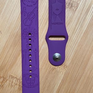 May include: Purple silicone Apple Watch band with a design featuring Wednesday Addams from the Netflix show "Wednesday". The band has a graphic of Wednesday Addams, the text "Wednesday", a cello, a spider web, a group of people, a person in a long coat, a sword, and the text "Nevermore Academy" with a crest.
