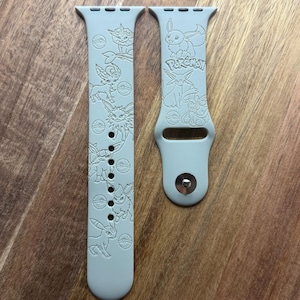 May include: Two light gray watch bands featuring engraved Pokémon characters and Poké Balls. One band has the word "Pokémon" engraved. The bands are designed for a smartwatch and are made of a flexible material.