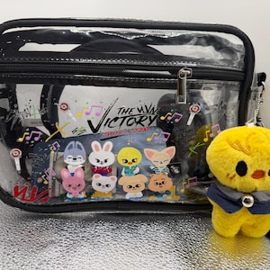 May include: A transparent crossbody bag with black edging and strap, displaying a colourful graphic of cartoon characters and the text "THE VICTORY". A yellow plush chick with a blue waistcoat hangs from the bag.