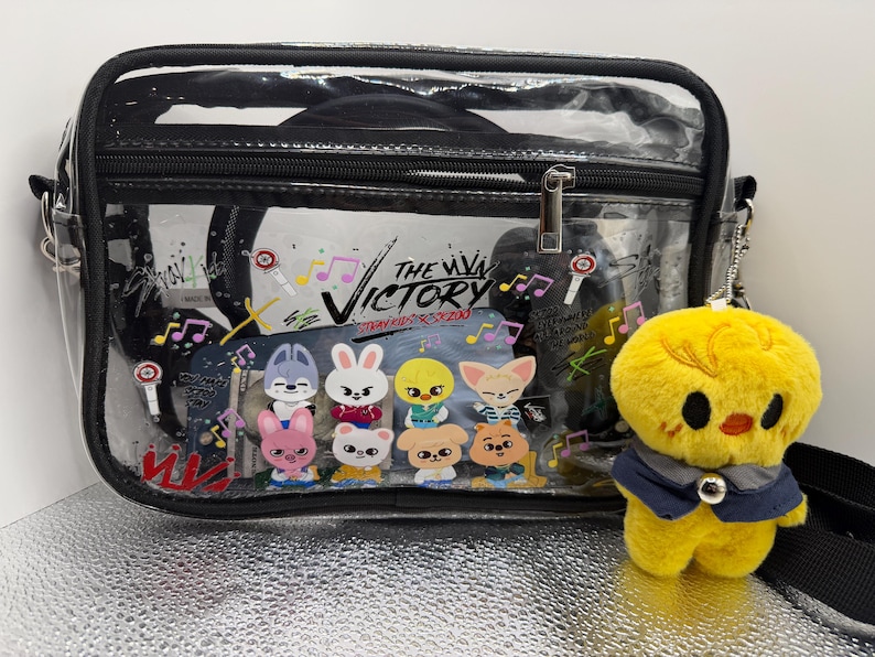 May include: A transparent crossbody bag with black edging and strap, displaying a colourful graphic of cartoon characters and the text "THE VICTORY". A yellow plush chick with a blue waistcoat hangs from the bag.