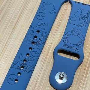 May include: Two blue watch straps with engraved cartoon character designs. One strap features multiple characters and a Poké Ball design. The other strap has the word "Snorlax" and a cartoon character design. The straps are made of a flexible material.