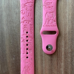 Puede incluir: Correa de reloj rosa con figuras de dibujos animados en relieve y los textos "BEH34" y "G-DRAGON". La correa tiene un pasador para la fijación. La correa está hecha de un material flexible.