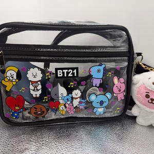 May include: A black and clear crossbody bag with a zipper closure and a black strap. The bag is decorated with colorful cartoon characters and the text "BT21". A white plush toy with a pink face hangs from the bag.