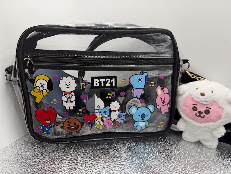 May include: A black and clear crossbody bag with a zip closure and a black strap. The bag is decorated with colourful cartoon characters and the text "BT21". A white plush toy with a pink face hangs from the bag.