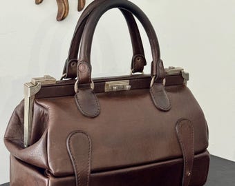 May include: A dark brown leather handbag featuring a structured rectangular base, a hinged metal frame closure, and two rounded top handles. The bag includes decorative leather tabs on the front and silver-toned metal hardware accents.