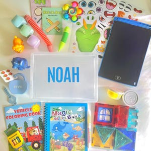 May include: A collection of children's toys and activities, including a clear plastic box with the name "NOAH" in blue, a coloring book, a drawing tablet, and various small toys. The items are arranged on a light-colored surface.