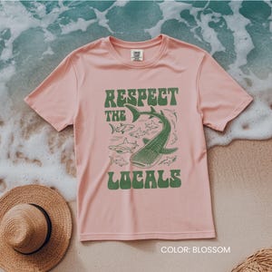Op de afbeelding: Lichtroze t-shirt met de groene tekst "Respect the Locals" en een afbeelding van een walvishaai en andere haaien. Het t-shirt heeft een ronde hals en is gemaakt van een zachte stof.