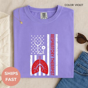 May include: Violet t-shirt with a graphic design featuring a white stethoscope, stars, and stripes, along with a red illustration of lungs and the words "Respiratory Therapist". The shirt is made of a soft material.