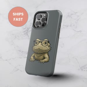 Op de afbeelding: Een grijze telefoonhoes met een cartoonillustratie van een baardagaam met een bril. De hagedis is bruin en heeft een stekelige baard. De hoes heeft een glanzende afwerking en een uitsparing voor de camera. De woorden "SHIPS FAST" staan in een perzikkleurige cirkel.