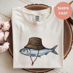 Op de afbeelding: Crèmekleurig T-shirt met een cartoonillustratie van een vis met een bruine vissershoed. De vis is blauw en zilver met een gedetailleerd ontwerp. Het shirt heeft een ronde hals en de woorden "SHIPS FAST" in een cirkel.