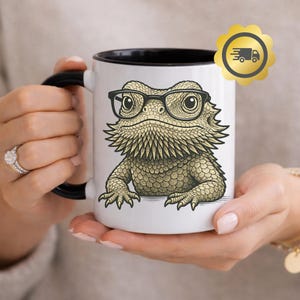 May include: White ceramic mug with a black interior and handle, featuring a detailed illustration of a bearded dragon wearing glasses. The lizard is in shades of tan and green. The mug is held by a person.