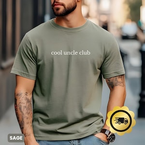 Op de afbeelding: Saliegroen t-shirt met de tekst "cool uncle club" in het wit. Het shirt heeft een ronde hals en lijkt van een zacht materiaal te zijn. De afbeelding bevat een bezorgicoon.