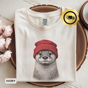 Op de afbeelding: Ivoor kleurig t-shirt met een gedetailleerde illustratie van een otter met een rode muts. Het shirt heeft een ronde hals en is gemaakt van zacht materiaal. Het merklabel is zichtbaar bovenaan. Het woord "IVORY" staat onderaan gedrukt.