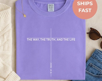 May include: A lilac-colored t-shirt with the text "THE WAY, THE TRUTH, AND THE LIFE" and "JOHN 14:6" printed in white. The shirt has a crew neck and short sleeves. A gold necklace and sunglasses are visible.