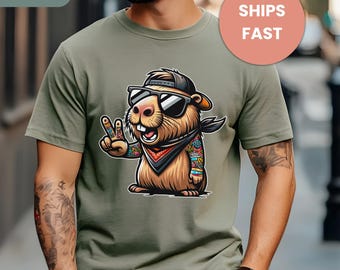May include: Sage green t-shirt featuring a cartoon groundhog wearing sunglasses, a bandana, and a cap, giving a peace sign. The groundhog has tattoos on its arm. The shirt has the word "SAGE" in the upper left corner.