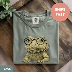 May include: Sage green t-shirt featuring a detailed illustration of a bearded dragon wearing round black glasses. The graphic is in shades of tan and gold. The shirt has a crew neck and short sleeves. The text "SHIPS FAST" is in a peach circle.