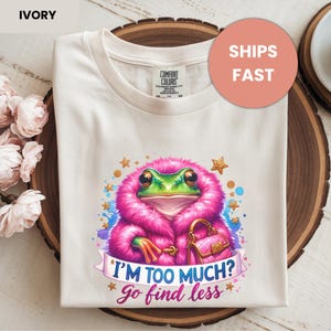 Op de afbeelding: Ivoor kleurig T-shirt met een grillige kikker illustratie. De kikker draagt een roze, pluizige jas en houdt een roze handtas vast, met de tekst "I'M TOO MUCH? Go find less". Het shirt is op een houten oppervlak gevouwen.