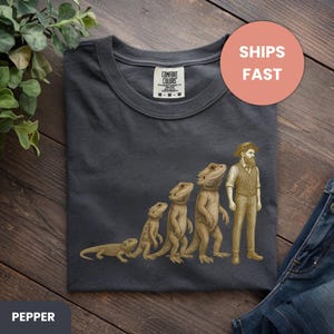 Op de afbeelding: Pepperkleurig t-shirt met een grappige afbeelding van de evolutie van een baardagaam naar een man. Het ontwerp toont een reeks hagedisachtige figuren die veranderen in een man met een hoed en vest. Met Comfort Colors label.