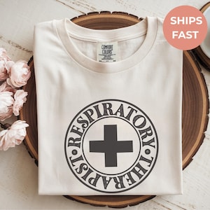 Puede incluir: Camiseta color crema con cuello redondo, con un diseño circular negro con las palabras "Respiratory Therapist" alrededor de un símbolo de cruz. La camiseta está doblada sobre una superficie de madera.