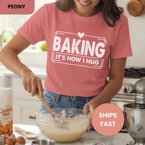 May include: A peony-colored t-shirt with the text "BAKING IT'S HOW I HUG" in a white rectangular design. The image shows a person mixing ingredients in a glass bowl with a wooden spoon. A bag of flour and a mixer are also visible.