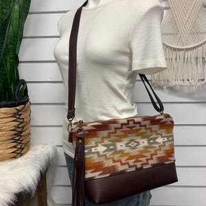 Peut inclure: Sac bandoulière avec un motif d'inspiration sud-ouest dans les tons marron, orange et crème. Le bas du sac est marron foncé et il est doté d'une longue bandoulière réglable. Une longue gland en cuir pend sur le côté.