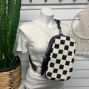 May include: A black and white checkered crossbody bag with a black leather strap and tassel. The bag is worn over a white t-shirt. A woven basket with a plant is in the background.