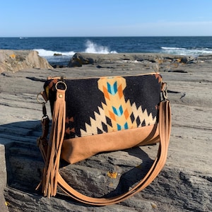 May include: A crossbody bag with a black patterned top and tan leather bottom. The bag features a geometric design in orange, blue, and tan. It has a long tan strap and a tassel. The bag is sitting on a rocky surface near the ocean.