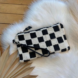 May include: A rectangular clutch bag featuring a black and white checkered pattern made of woven fabric. The bag includes a black zipper closure along the top edge and a detachable black wrist strap attached to a silver-toned metal clasp on the side.