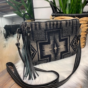 Puede incluir: Un bolso cruzado con estampado negro y beige con una correa y borla de cuero negro. El bolso presenta un diseño geométrico y una cremallera plateada. La correa tiene costuras blancas.