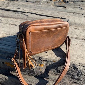 May include: A brown leather crossbody bag with a rectangular shape. The bag features a front pocket, a zipper closure, and a long adjustable strap. A decorative tassel hangs from the side. The bag is made of a textured material.