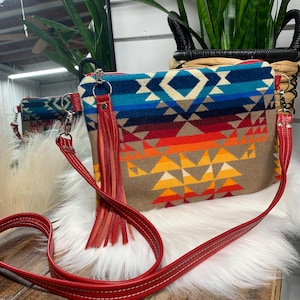 Puede incluir: Un bolso bandolera rectangular con un colorido estampado geométrico en tonos azules, naranjas y beige. El bolso tiene una correa y borla de cuero rojo, y una cremallera plateada. El bolso está sobre una superficie blanca y esponjosa.