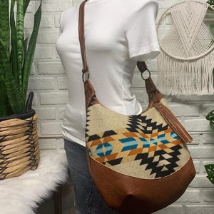 Peut inclure: Un sac à bandoulière de style hobo avec un panneau en tissu crème orné d'un motif géométrique noir, beige et bleu clair. Le sac présente des finitions en similicuir marron, une bandoulière assortie et un pompon décoratif sur un anneau en métal.
